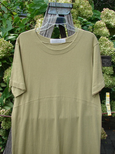 1998 Botanicals Calla Dress in Echinacea Seed, size 0, shown in a muted olive tone with a smooth, draped knit body and short sleeves. The dress hangs on a clear hanger against greenery, with soft natural light revealing its simple round neckline and gently curved seam.