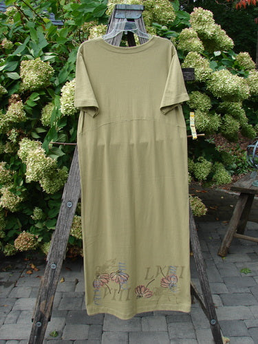 1998 Botanicals Calla Dress Echinacea Seed Size 0 in muted seed green, a straight short-sleeve dress with a simple crew neckline and soft drape. Floral embroidery and faint lettering decorate the lower hem, while the garment hangs on a wooden ladder amid dense garden greenery.