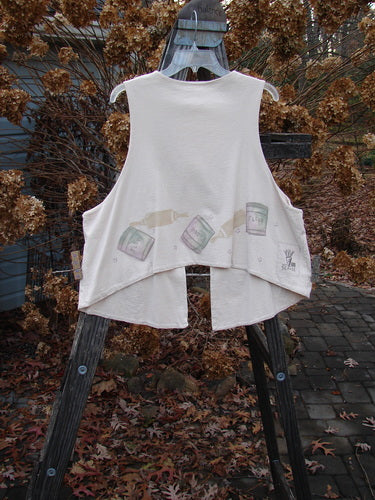Image: A white shirt with a painted design on it, hanging on a clothesline outdoors. 

Alt Text: 1999 The Tab Vest Baker Tea Dye Size 2: A white shirt with a baking-themed painted design, hanging on a clothesline outdoors.