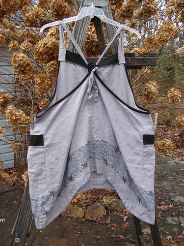 Barclay PMU Patched Linen Contrast Apron Jumper Echinacea Black Dusk Size 1: A grey dress on a wooden pole, with versatile shoulder straps, a botanicals patch, and wrap around drop pockets.