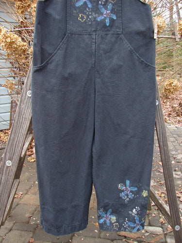 A black Parachute Overall with oversized front bib pocket, deep side pockets, adjustable shoulder straps, metal button side adjustments, wide swingy lowers, drawcord back, and a floral theme paint. Size 0.