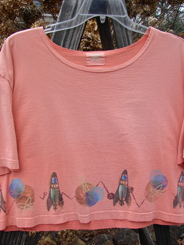 2000 Short Sleeved Crop Tee Rocket Coral Size 1: A pink shirt with rocket designs on it, featuring a wider flatly finished neckline and a super wide crop box shape.