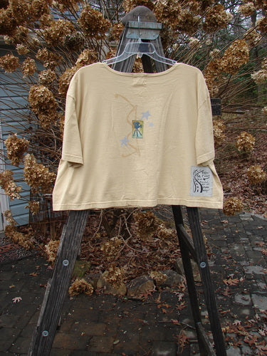2000 Short Sleeved Crop Tee Flamingo Plantain Size 2: A shirt on a swinger with a tan shirt on a wooden stand. Close-up of a plant and a pile of leaves on a wooden stand. Close-up of a swinger and a wooden plank with a white object on it. Close-up of a stone floor.