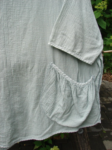 Barclay Gauze Round Bottom Tie Back Pocket Top Unpainted Grass Size 2: A light-gray textured cotton/linen smock with a loose fit, featuring a gathered patch pocket, hung outdoors amidst green leaves and paving stones.