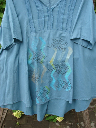 Barclay Batiste Flutter Bib Tie Back Dress Honeycomb Tealen Size 1 is showcased draped on a wooden bench, highlighting its square yoke with pintucked detailing and vibrant fish-scale print.