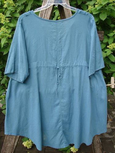Barclay Batiste Flutter Bib Tie Back Dress Honeycomb Tealen Size 1: A loose-fitting tunic with wide sleeves, a rounded neckline, and a back tie, displayed outdoors against a wooden support with foliage.