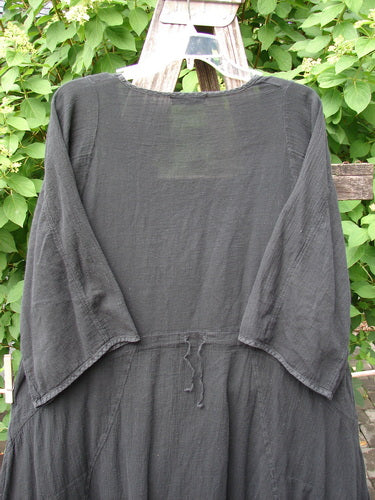 Barclay Gauze Urchin Bell Dress Unpainted Black Size 1, a loose-fitting dress with ¾-length sleeves and a waist tie, is displayed outdoors on a wooden hanger, highlighting its elegant, airy design.