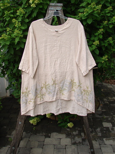 Barclay Linen Hi Low Tunic Top Bamboo Border Plantain Size 2 displayed outdoors on a wooden easel, featuring a round neckline, elbow-length sleeves, and sage-green leaf motifs along the hem.
