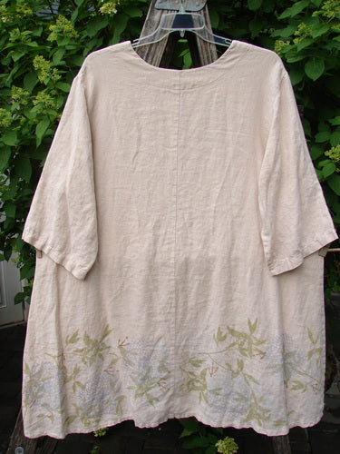 Barclay Linen Hi Low Tunic Top Bamboo Border Plantain Size 2, featuring a soft cream hue, round neckline, elbow-length sleeves, and nature-inspired leaf and branch motifs along the hem, on a wooden hanger outdoors.