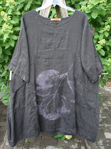 Barclay Linen Sectional Urchin Dress Leaf Black Size 1 hangs outdoors, showcasing its elbow-length sleeves, empire waist, side pockets, and large purple leaf graphic on a dark gray tunic.