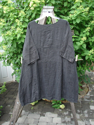Barclay Linen Sectional Urchin Dress Leaf Black Size 1: A loose-fitting black dress with three-quarter sleeves, featuring an empire waist, varying hemline, and double side pockets, displayed on a rustic wooden ladder.