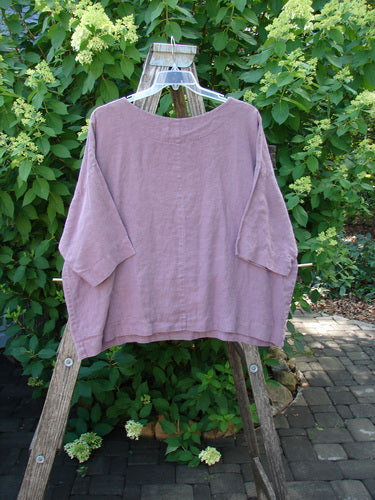 Barclay Linen Lantern Boxy Unpainted Dark Mauve Size 1: A loose-fitting mauve linen top with drop shoulders and a rounded neckline, displayed on a hanger against a leafy garden backdrop.