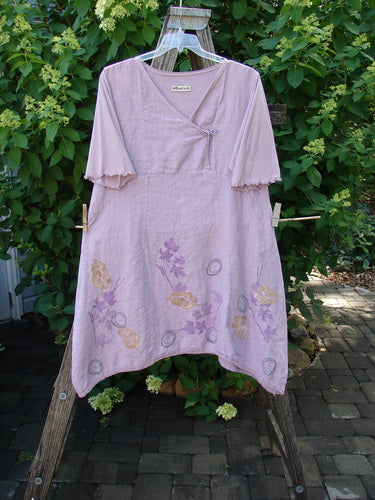 Barclay Linen Lace Blooming Tunic Dress Leaf Vine Lavender Size 0 hangs on a wooden ladder, showcasing its elbow-length sleeves, crossover neckline, and handkerchief hem with floral and circular motifs.
