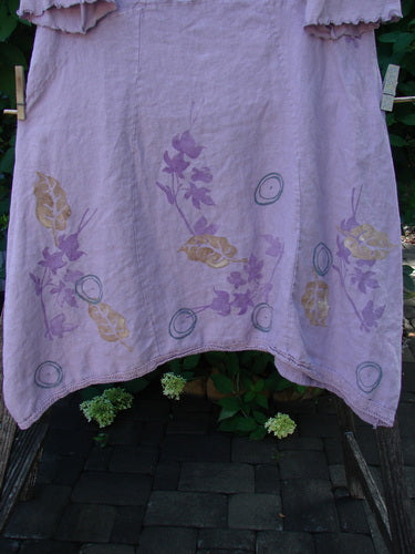 Barclay Linen Lace Blooming Tunic Dress Leaf Vine Lavender Size 0, featuring a cross-over neckline, floral motifs, and a lace-trimmed hem, hanging on a wooden rack with green foliage in the background.