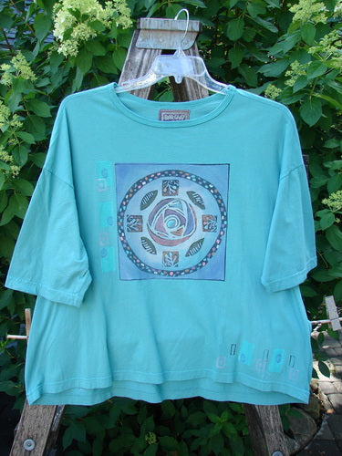 Barclay Short Sleeved Crop Box Tee Center Rose Aqua Size 3, featuring a mandala-style rose graphic, hangs on a clear hanger against leafy shrubs, showcasing its oversized boxy shape and soft, airy organic cotton.
