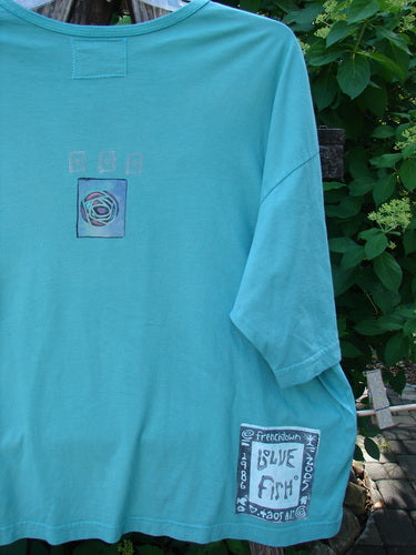 Barclay Short Sleeved Crop Box Tee Center Rose Aqua Size 3 hangs against foliage, featuring a circular motif on the chest and Blue Fish print at the hem, embodying Bluefishfinder.com's vintage aesthetic.
