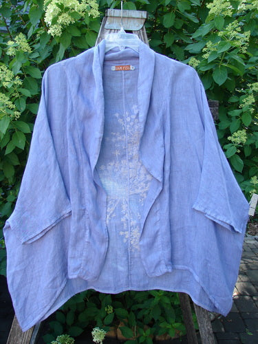 Barclay Linen Open Front Cheerful Cardigan Jacket Rear Starburst Iris Size 1, features a muted lavender tone, dolman sleeves, draped panels, and floral embroidery, hanging against green foliage.