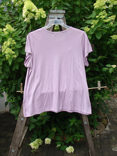 Barclay Cap Sleeved Tee Unpainted Lavender Size 2, displayed on a clear hanger, draped over a wooden structure with a lush greenery backdrop, reflecting Bluefishfinder.com's vintage, expressive clothing ethos.