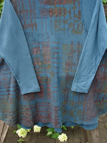 Barclay Thermal Monte Verde Vented Tunic Grid Blend Tealen OSFA hangs against a wooden backdrop, showcasing its slate blue fabric with rust-orange and green botanical patterns, featuring side vents and ribbed sleeve accents.