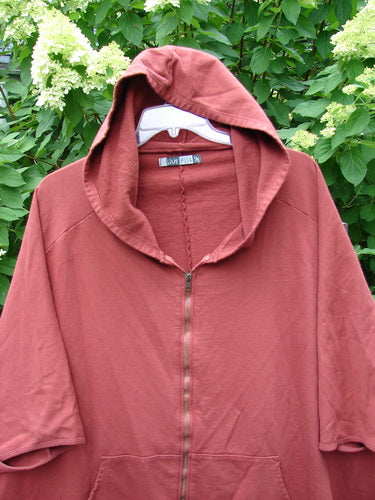 Barclay PMU Patched Hooded Postcard Zipper Jacket Vest in Deep Brick hangs on a white hanger, showcasing its short sleeves, full zipper front, and unique rear patch against lush greenery.