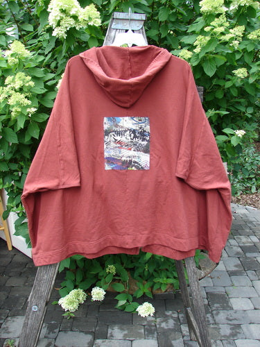 Barclay PMU Patched Hooded Postcard Zipper Jacket Vest in Deep Brick OSFA draped on a garden trellis, showcasing a graphic rear patch with abstract patterns against a lush, leafy backdrop.