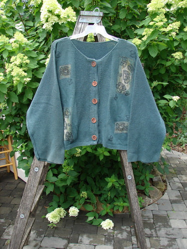 1994 Panel Cardigan Sweater Magic Tea Party Nightshade OSFA: A loose-fitting, forest-green cardigan with wooden buttons, subtle patchwork, and fold-over belled sleeves, hanging on a wooden easel against a garden backdrop.