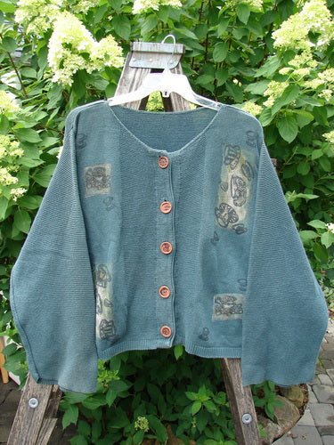 1994 Panel Cardigan Sweater Magic Tea Party Nightshade OSFA, showcasing a teal-green knit with wooden buttons, floral motifs, and bell sleeves, draped over a wooden ladder against lush greenery.