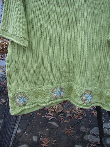 1999 Spring Pullover Tunic Sweater Kiwi Size 2: A green sweater with embroidery and blue flowers, featuring varied textures and knits, drop shoulders, and rolled sleeves and hemline.