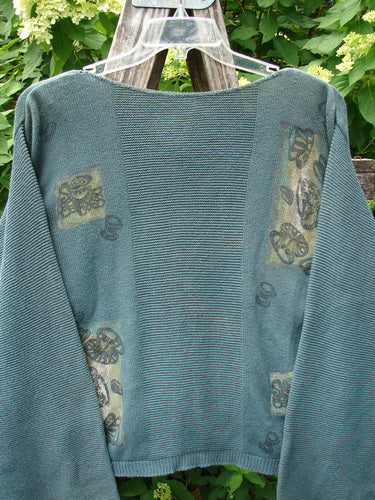 1994 Panel Cardigan Sweater Magic Tea Party Nightshade OSFA: Teal-green cardigan with stamped wheel motifs, fold-over belled sleeves, and boxy shape, draped on a wooden ladder amidst a floral backdrop.