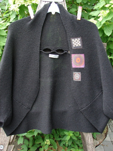 Barclay Patched Alpaca Rear Window Shrug Black OSFA, featuring a wide collar, colorful patches, open front, and draped design, displayed on a hanger with leafy foliage background.