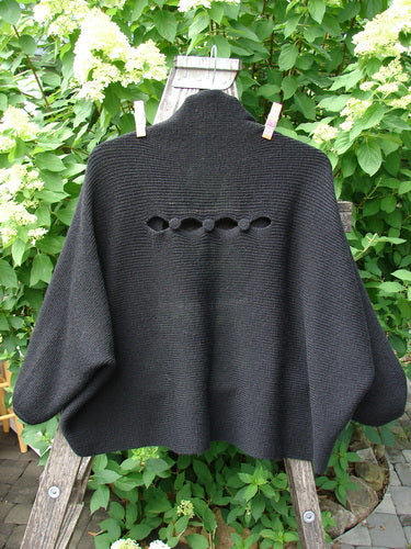 Barclay Patched Alpaca Rear Window Shrug Black OSFA displayed on a wooden ladder, showcasing its bat-wing style, oval eyelets, and unique texture, framed by lush outdoor greenery.