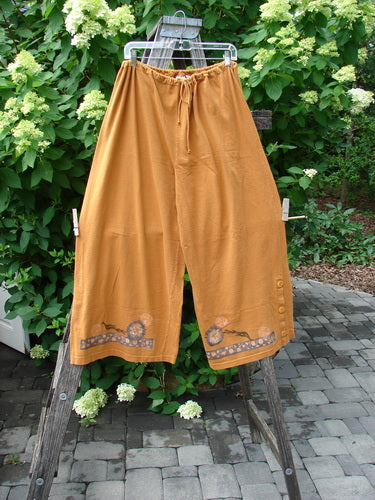 The 1994 Banjo Pant Dandelion Breeze Oro Size 2 features loose-fitting, culotte-style pants with wide legs, decorative hem prints, and side buttons, hanging outdoors against lush greenery.