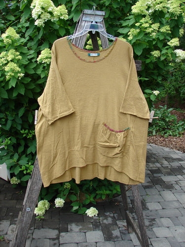 Barclay Gauze Round Bottom Tie Back Pocket Top Ditsy Daisy Golden Size 2, displayed on a rustic frame amidst greenery, showcases its loose fit, elbow-length sleeves, and front patch pocket.