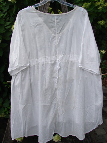 Barclay Batiste Flutter Bib Tie Back Dress, a loose-fitting, white blouse with a scooped neckline, three-quarter sleeves, and waist tie, hanging gracefully on a clear hanger amidst garden foliage.