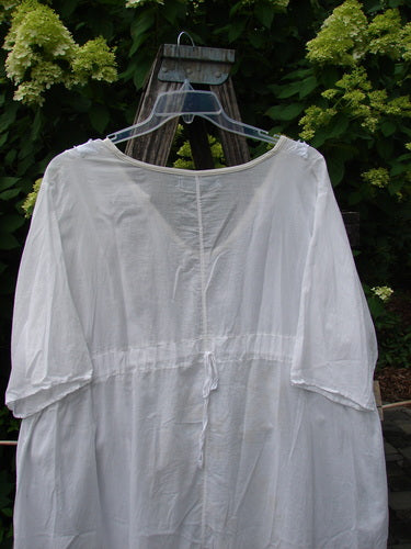 Barclay Batiste Flutter Bib Tie Back Dress in Triangular Geo White, Size 2, displayed on a clear hanger over a wooden step ladder, with leafy green shrubs in the background.
