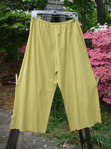 A pair of Barclay Crop Tiny Tab Pants in Sunshine, size 2, on a clothesline. Made from medium weight cotton lycra, these pants feature a full elastic waistband, lettuce-edged swinging lowers, and a fun unpainted layering piece. Waist: 30-40, Hips: 54, Inseam: 23, Length: 35.