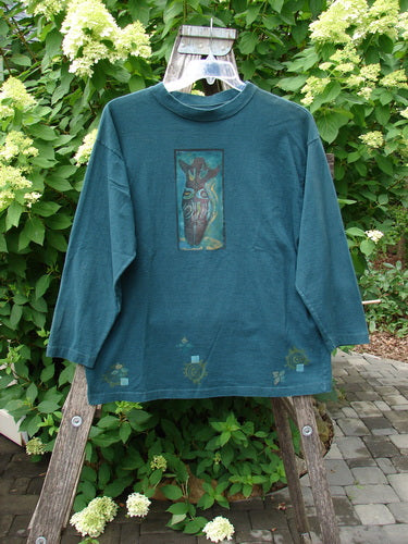 1996 Long Sleeved Tee Primitive Mask Viridian Size 1 displayed on a wooden ladder, featuring a bird graphic on the chest and decorative motifs near the hem, amidst a lush garden setting.
