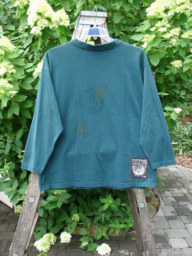 1996 Long Sleeved Tee Primitive Mask Viridian Size 1, draped over a wooden stepladder, showcases drop shoulders, thick ribbed neckline, cozy long sleeves, and a signature Blue Fish patch amidst lush foliage.