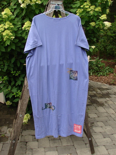 1997 Aquavella Dress Celtic Trio Skylark Size 2: A loose lavender T-shirt dress with abstract patches, hanging on a wooden rack outdoors amidst lush greenery, showcasing vintage Blue Fish Clothing's creative, expressive style.