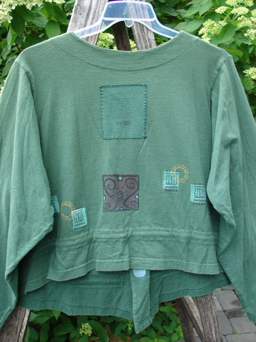 1997 Treehouse Jacket Greek Window Verdigris Size 2: A green cropped jacket with patch-style decorations and mug motifs, featuring a double-paneled V-neck, button line, and adjustable flounce tie, photographed outdoors.