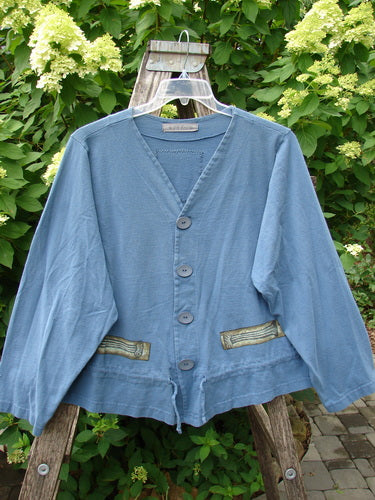 1997 Treehouse Jacket Greek Column Blueprint Size 2, a slate-blue cardigan with a V-neck, four buttons, patch pockets, and decorative trim, elegantly draped on a rustic wooden ladder amidst lush foliage.