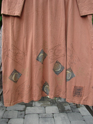 The 1998 Mandolin Dress Lucky Horseshoe Rockwood Size 2 features a loose-fitting, long-sleeved silhouette with block-print motifs along the hem, hanging on a wooden rail above a textured stone floor.