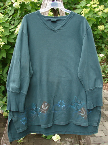 Barclay Interlock V Neck Vented Tunic Top Celtic Tumble Forest Teal Size 2; a loose-fit, long-sleeve pullover with a floral motif along the hem, displayed outdoors against a garden backdrop.