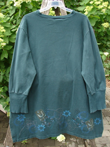 Barclay Interlock V Neck Vented Tunic Top Celtic Tumble Forest Teal Size 2, showcasing a ribbed neckline, vented sides, and geometric prints on the hem, hangs outdoors against leafy shrubs.