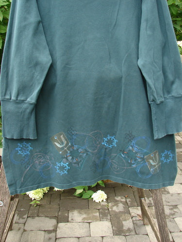 Barclay Interlock V Neck Vented Tunic Top Celtic Tumble Forest Teal Size 2, featuring a thicker ribbed neckline, snowflake-like hemline motifs, and ribbed lower sleeves, draped on an outdoor wooden bench.