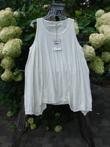 Magnolia Pearl NWT Cotton Paz Tank White OSFA displayed on a hanger, showcasing its raw edges, A-line flare, and hand-distressed texture, set against a lush green and floral backdrop.