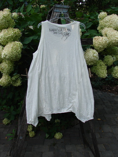 Magnolia Pearl NWT Cotton Paz Tank White OSFA features a sleeveless, tunic-length design with MAGNOLIA PEARL printed on the chest, displayed outdoors with lush foliage and hydrangea-style blossoms in the background.