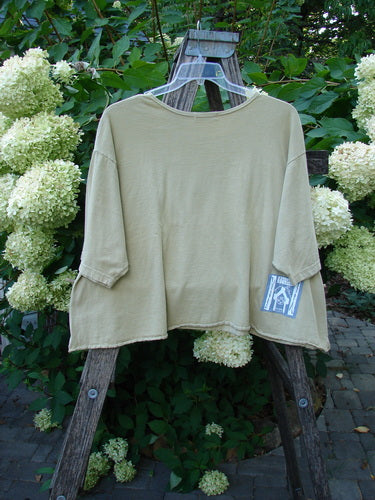 Barclay PMU Patched Short Sleeved Crop A Lined Tee Honey Size 2 displayed on a wooden A-frame, adorned with a front leaf-themed patch, amidst lush garden foliage and hydrangeas.