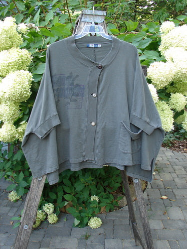 Barclay Slub Dolman Shirttail Pocket Jacket Fern Green Army Size 0 on wooden stand, featuring a round neckline, button-front, dolman sleeves, patch pockets, shirttail hemline, and subtle chest graphic against a garden backdrop.