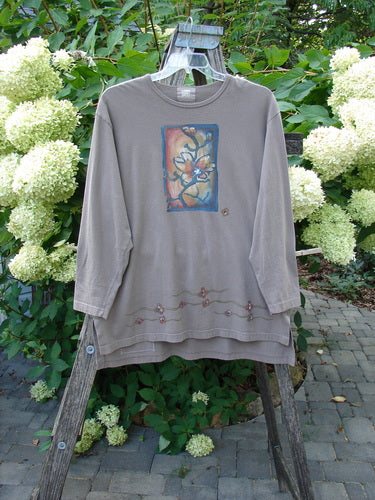1999 Long Sleeved Vented Tee Orchid Sprig Shale Size 1, displayed on a wooden ladder with a watercolor floral motif on the chest and decorative hem, set against a garden backdrop.